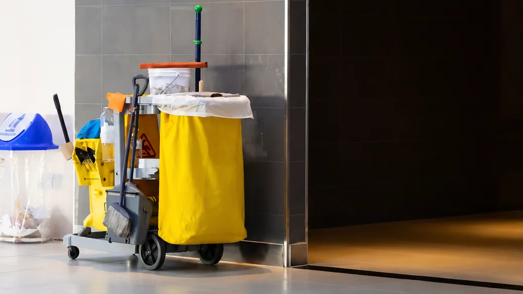 Janitorial cart with a mop, broom, and trash liner parked against a tiled wall in a commercial building lobby
